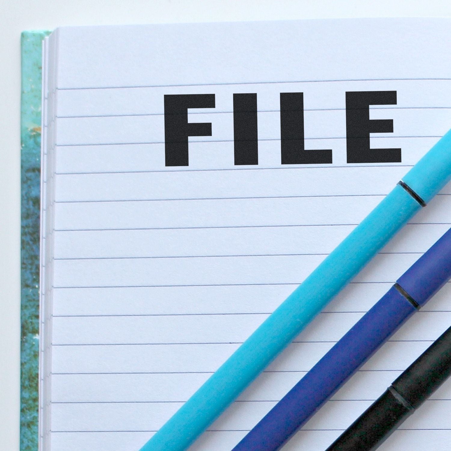 Large Pre-Inked File Stamp imprint on a lined notebook page with two pens, one blue and one black, placed diagonally across the page.