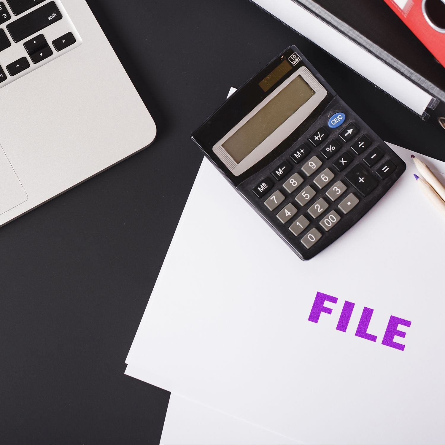 A Large Pre-Inked File Stamp marks a stack of papers labeled FILE next to a calculator, pencil, and part of a laptop on a black desk.