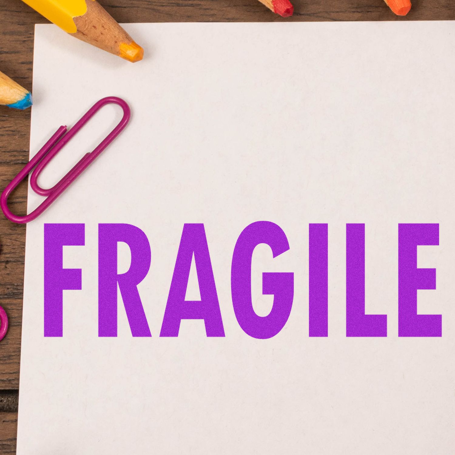 Large Pre-Inked Fragile Stamp in purple ink on white paper, surrounded by colorful pencils and a pink paperclip on a wooden surface.