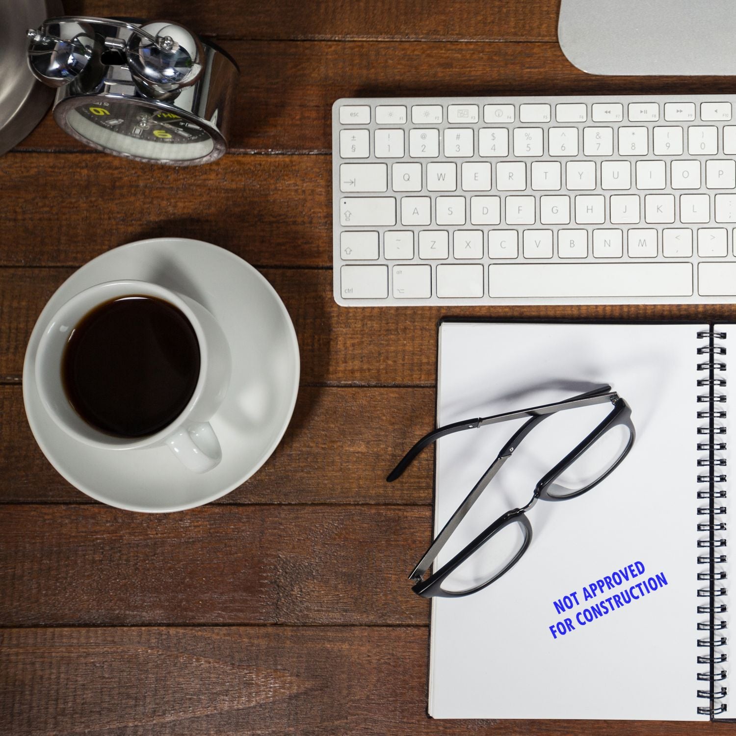 A desk with coffee, glasses, keyboard, and a notebook stamped with Not Approved For Construction using a Large Pre-Inked Not Approved For Construction Stamp.