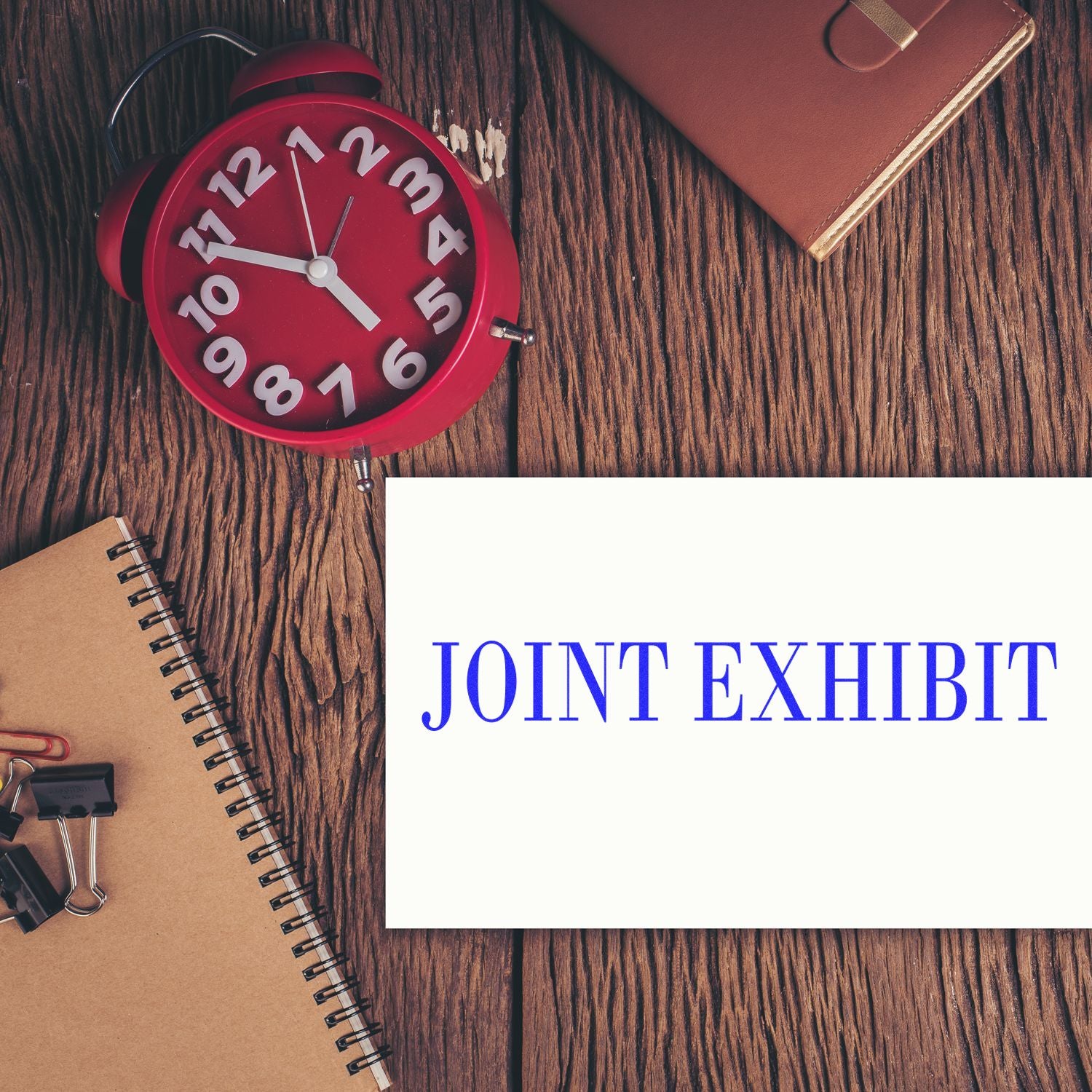 A Large Pre-Inked Joint Exhibit Stamp is placed on a wooden desk with a red clock, a notebook, and office supplies.