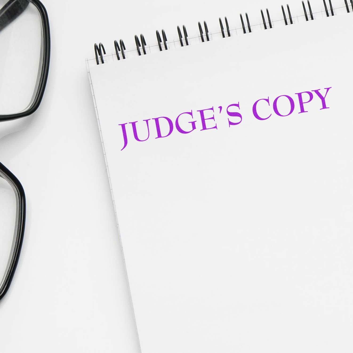 Large Pre-Inked Judge's Copy Stamp in purple ink on a white notepad, with black-rimmed glasses placed beside it.