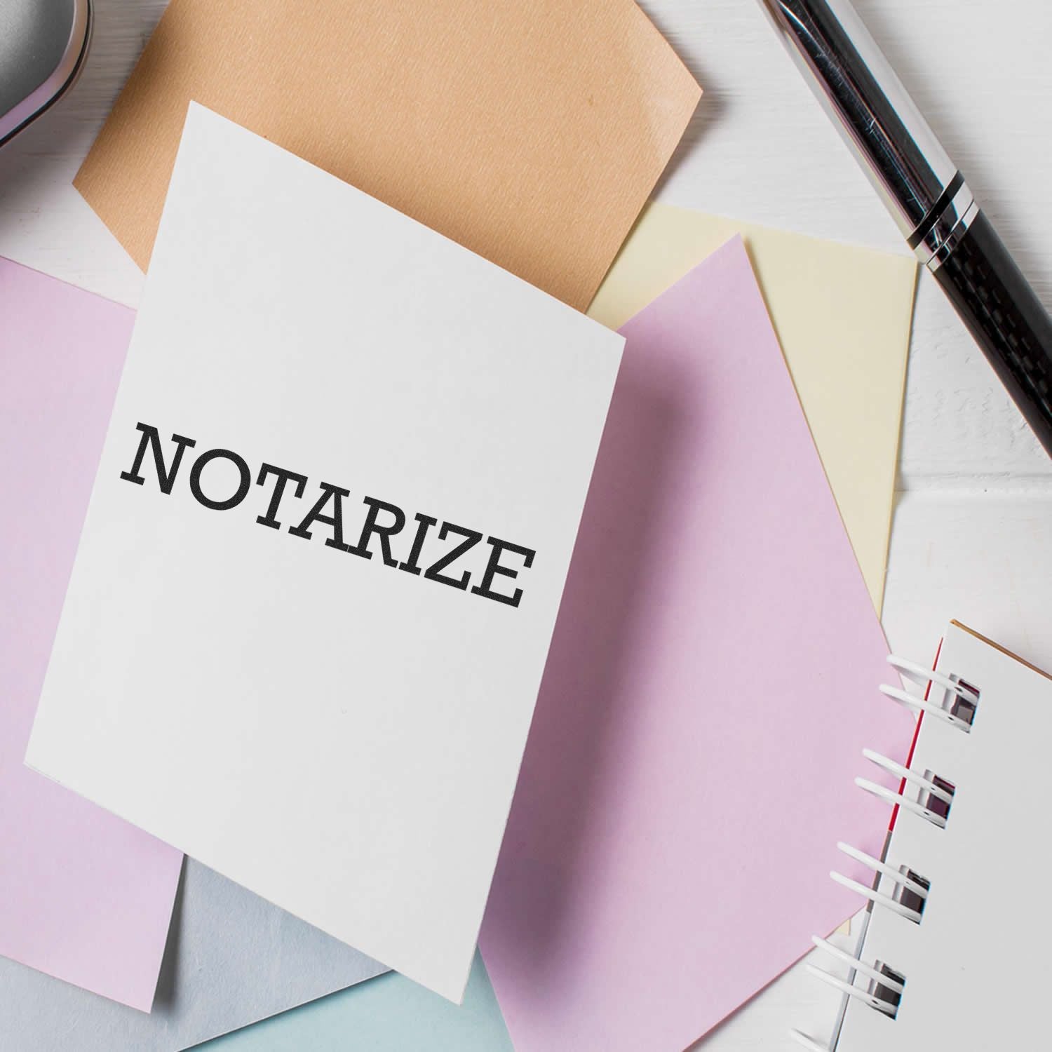 Large Pre-Inked Notarize Stamp on a white card surrounded by pastel-colored papers, a pen, and a notebook on a white surface.