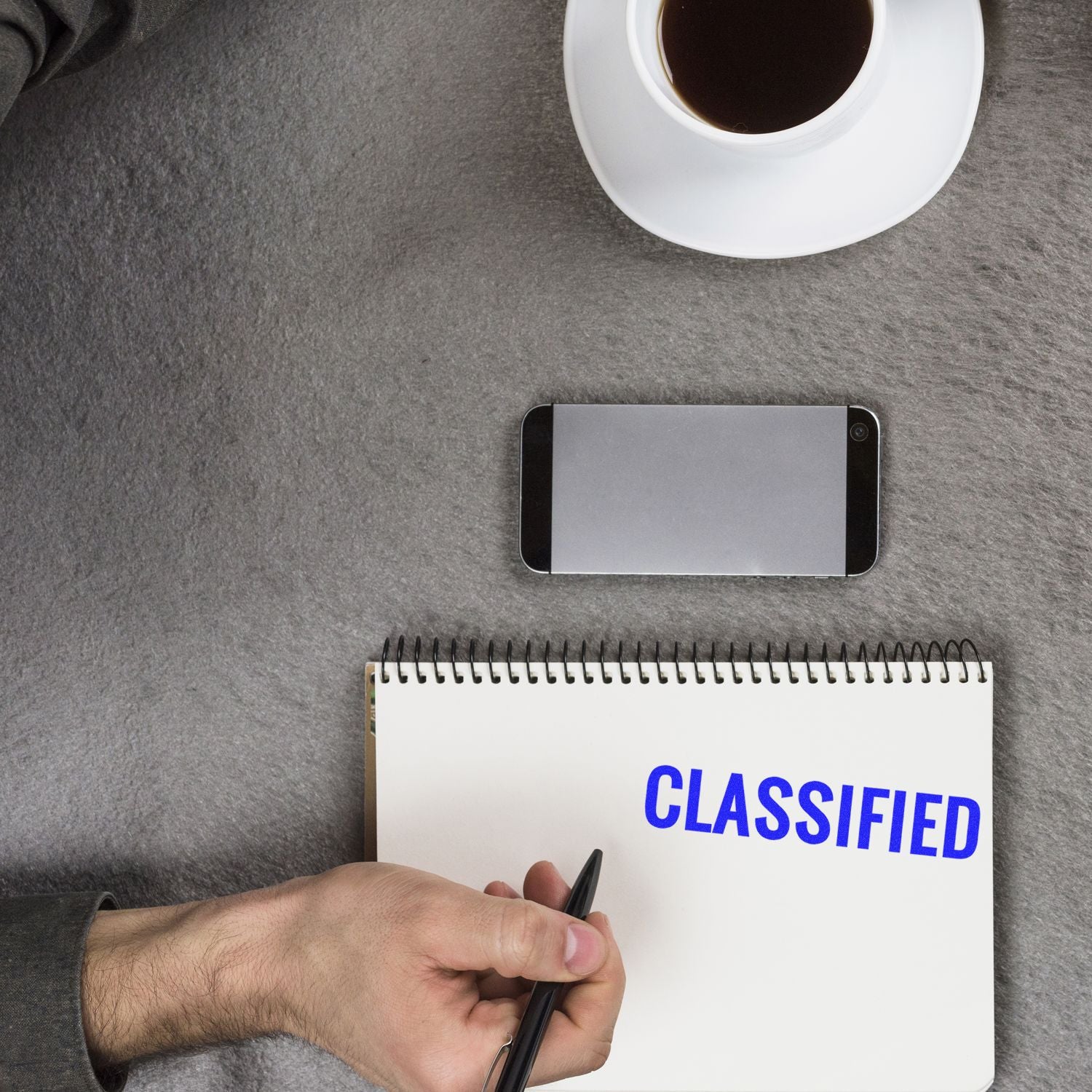 Hand holding a Large Pre-Inked Classified Stamp, stamping CLASSIFIED on a notepad, with a smartphone and coffee cup nearby on a gray surface.