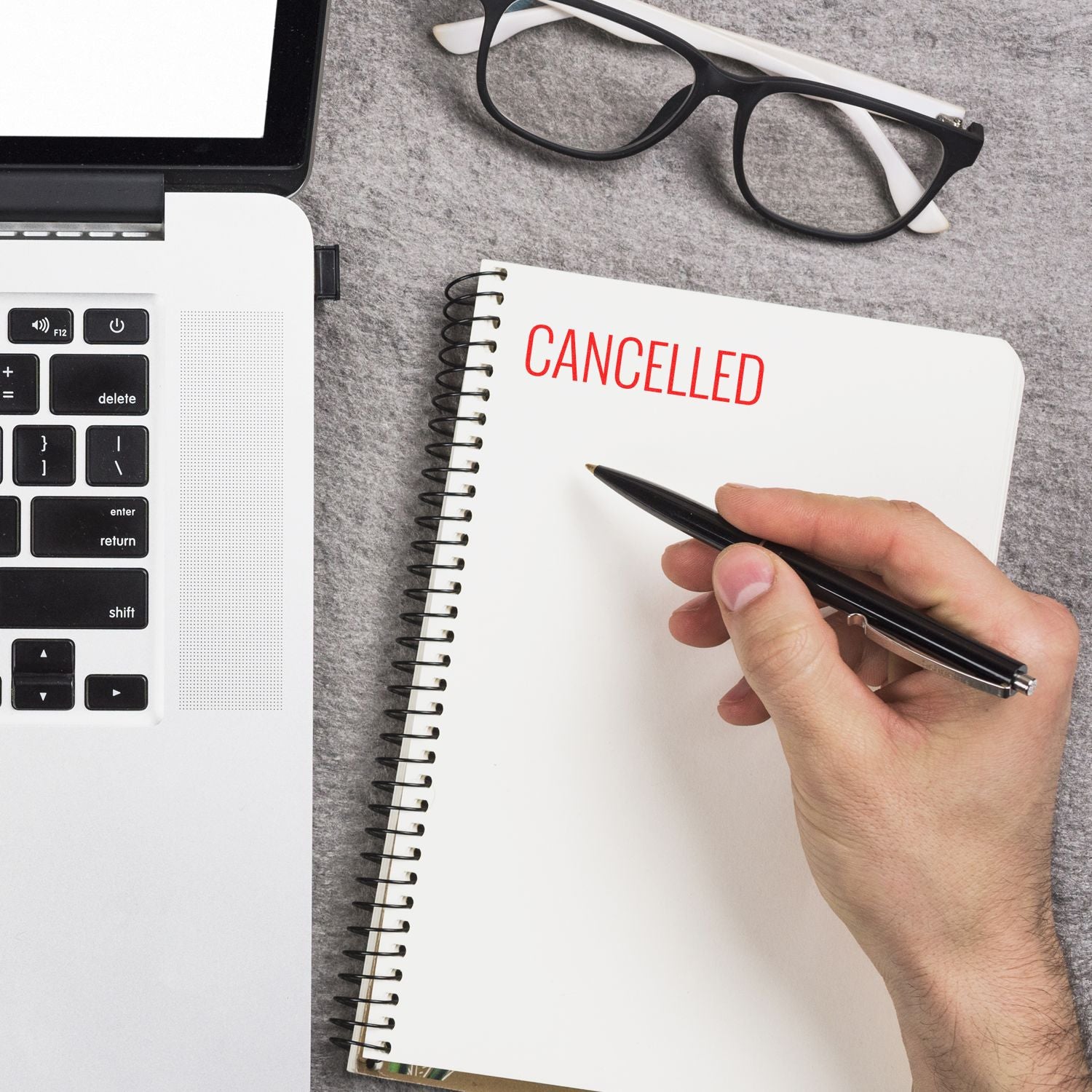 A hand holding a pen near a notebook with CANCELLED stamped in red using the Large Pre-Inked Narrow Font Cancelled Stamp, next to a laptop and glasses.