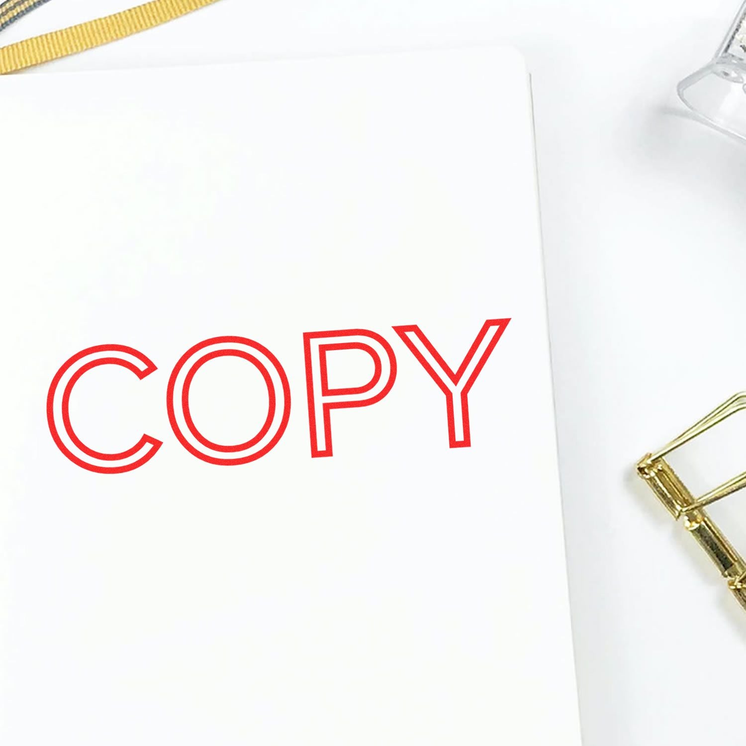 Large Pre-Inked Outline Font Copy Stamp in red ink on white paper, with a gold paperclip and a clear plastic item nearby.