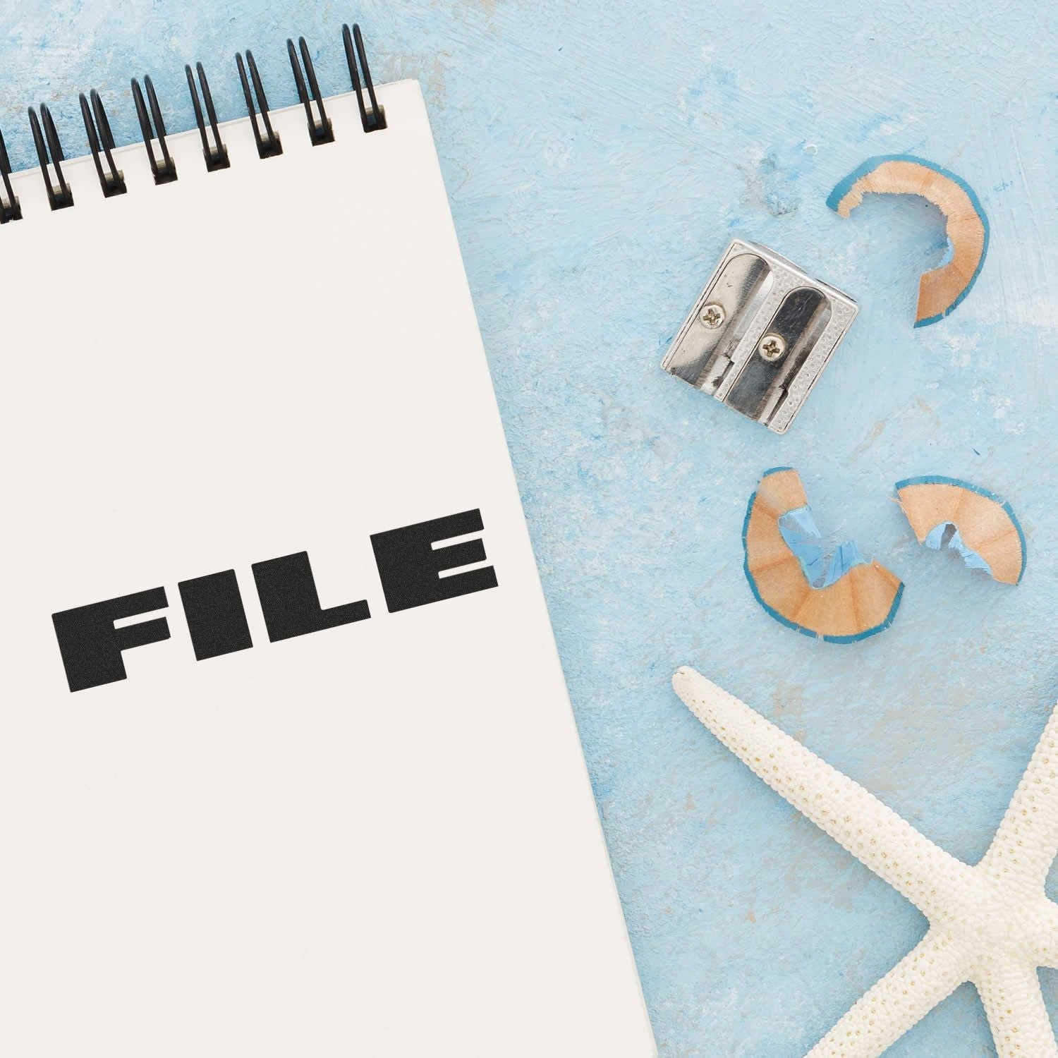 Large Pre-Inked Bold Font File Stamp on a spiral notebook, with a pencil sharpener, shavings, and a starfish on a blue surface.