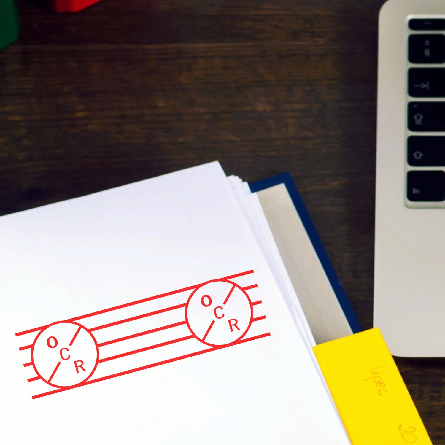 Large Pre-Inked OCR Stamp in red ink on a white paper, placed on a wooden desk next to a laptop and a yellow sticky note.