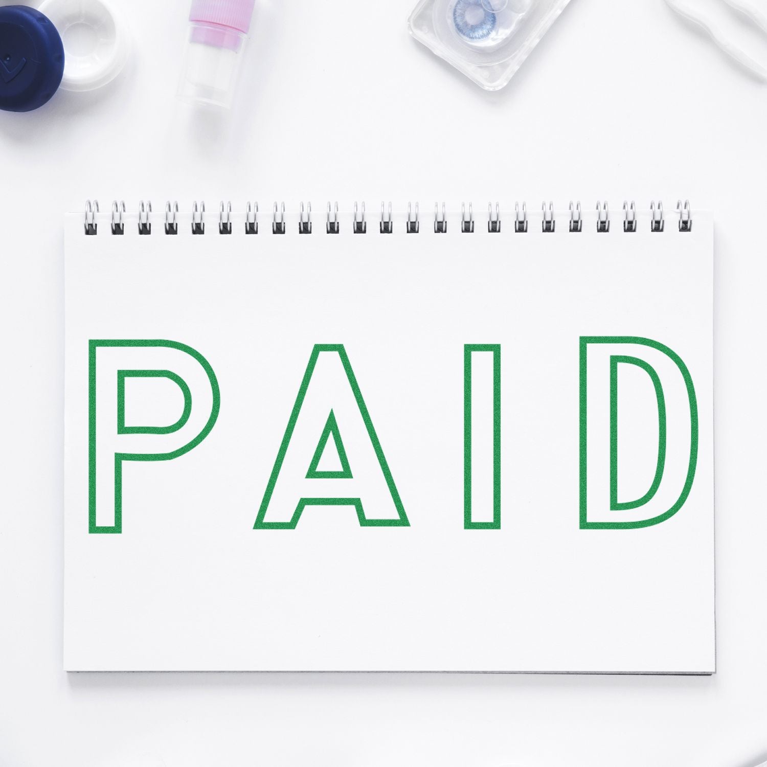 Large Pre-Inked Outline Paid Stamp in green ink on a white notepad, surrounded by office supplies on a white background.