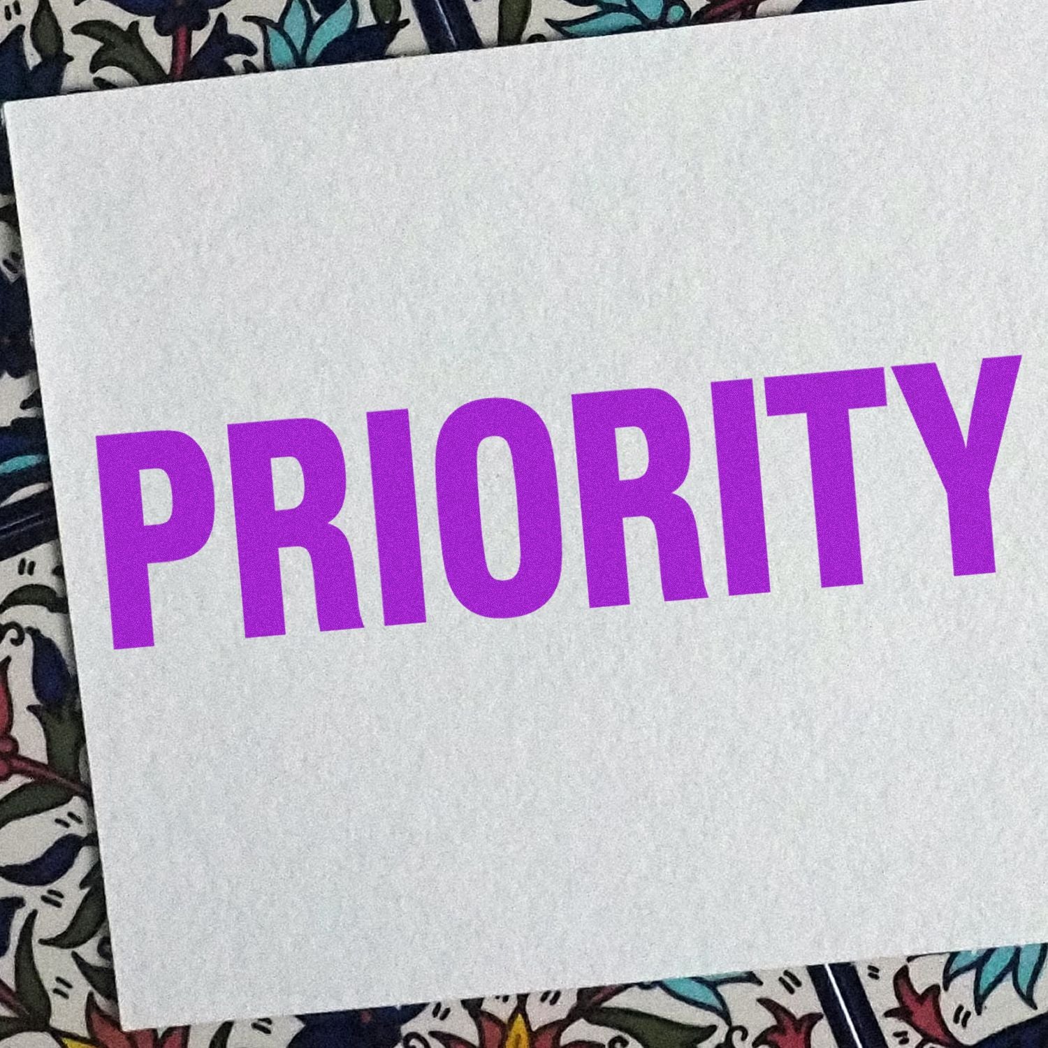 Large Pre-Inked Bold Priority Stamp in purple ink on white paper, placed on a colorful patterned background.