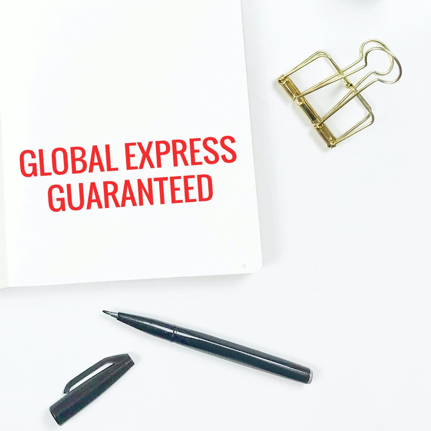 Large Pre-Inked Global Express Guaranteed Stamp on white paper, with a black pen, pen cap, and gold binder clip nearby.