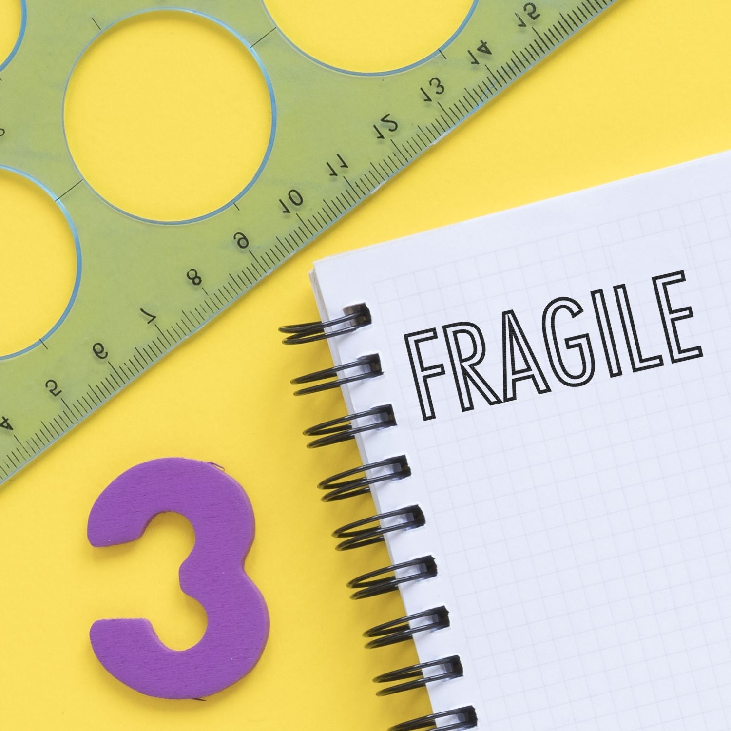 Large Pre-Inked Fragile Stamp on a spiral notebook with a green ruler and a purple number 3 on a yellow background.