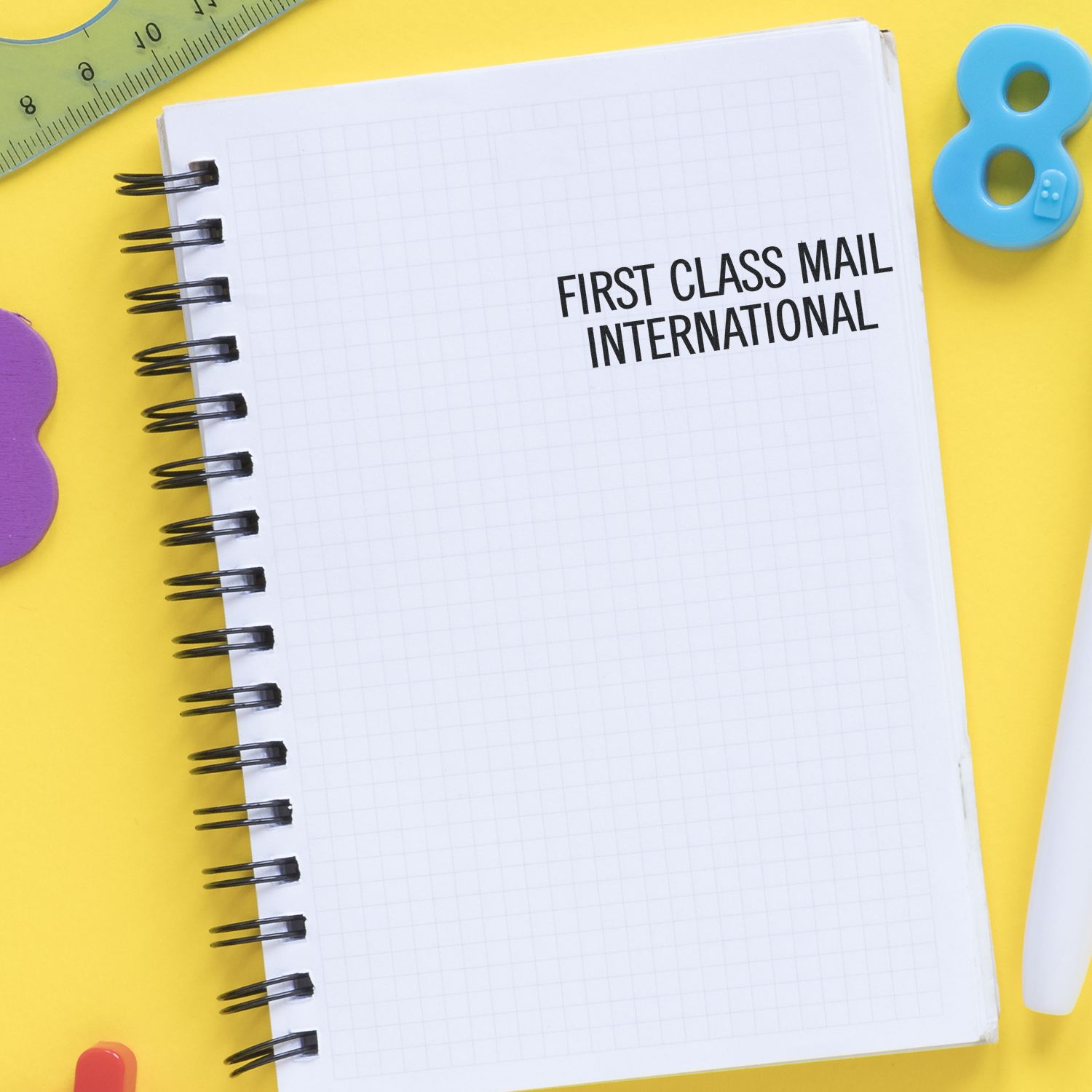 Large Pre-Inked First Class Mail International Stamp on a white notebook with grid paper, surrounded by colorful stationery on a yellow background.