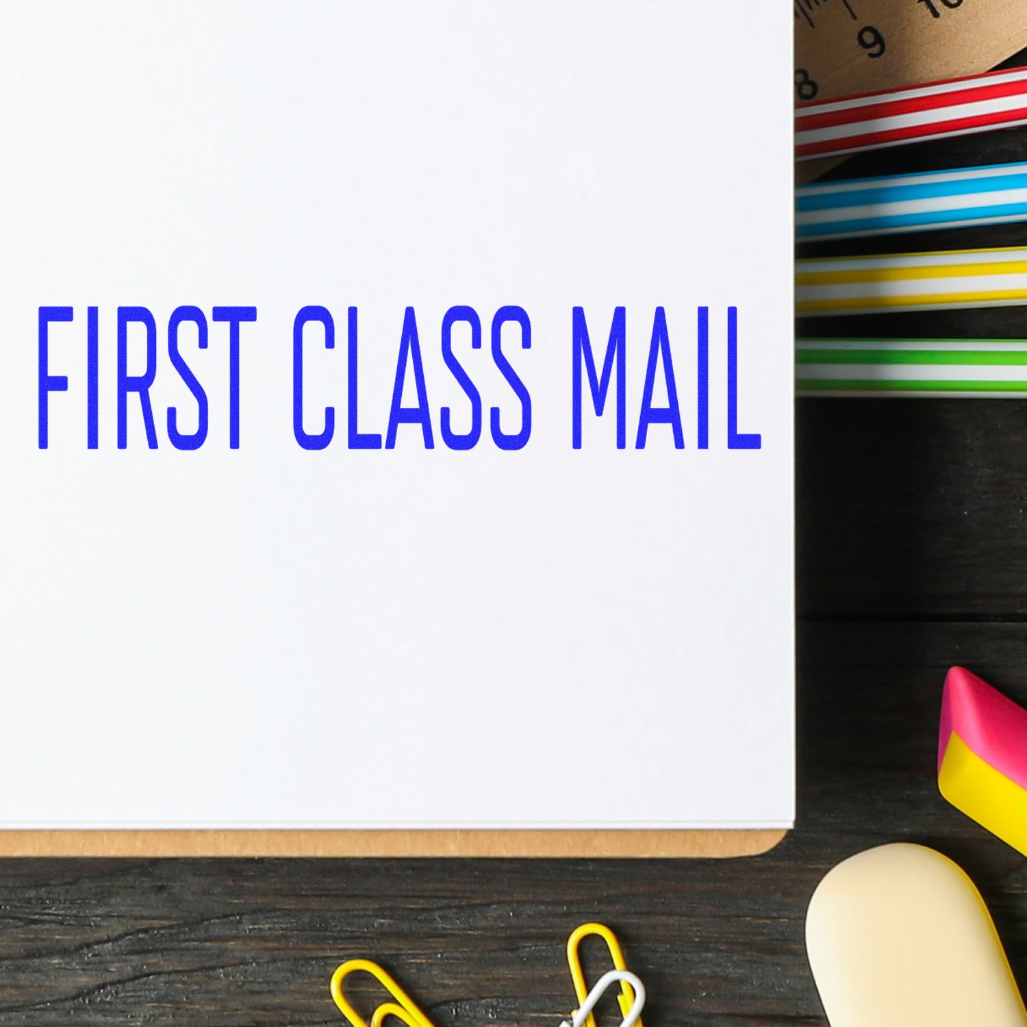 Large Pre-Inked Narrow Font First Class Mail Stamp imprint on white paper, surrounded by colorful office supplies on a dark wooden desk.