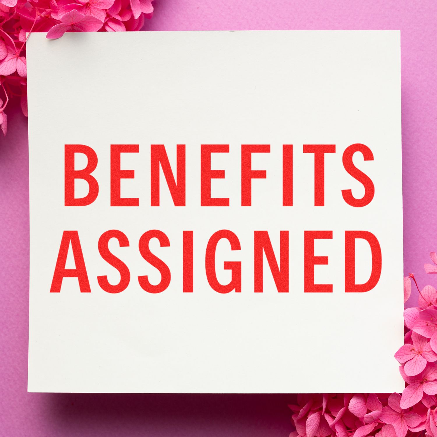 Large Pre-Inked Benefits Assigned Stamp in red ink on white paper, surrounded by pink flowers on a pink background.