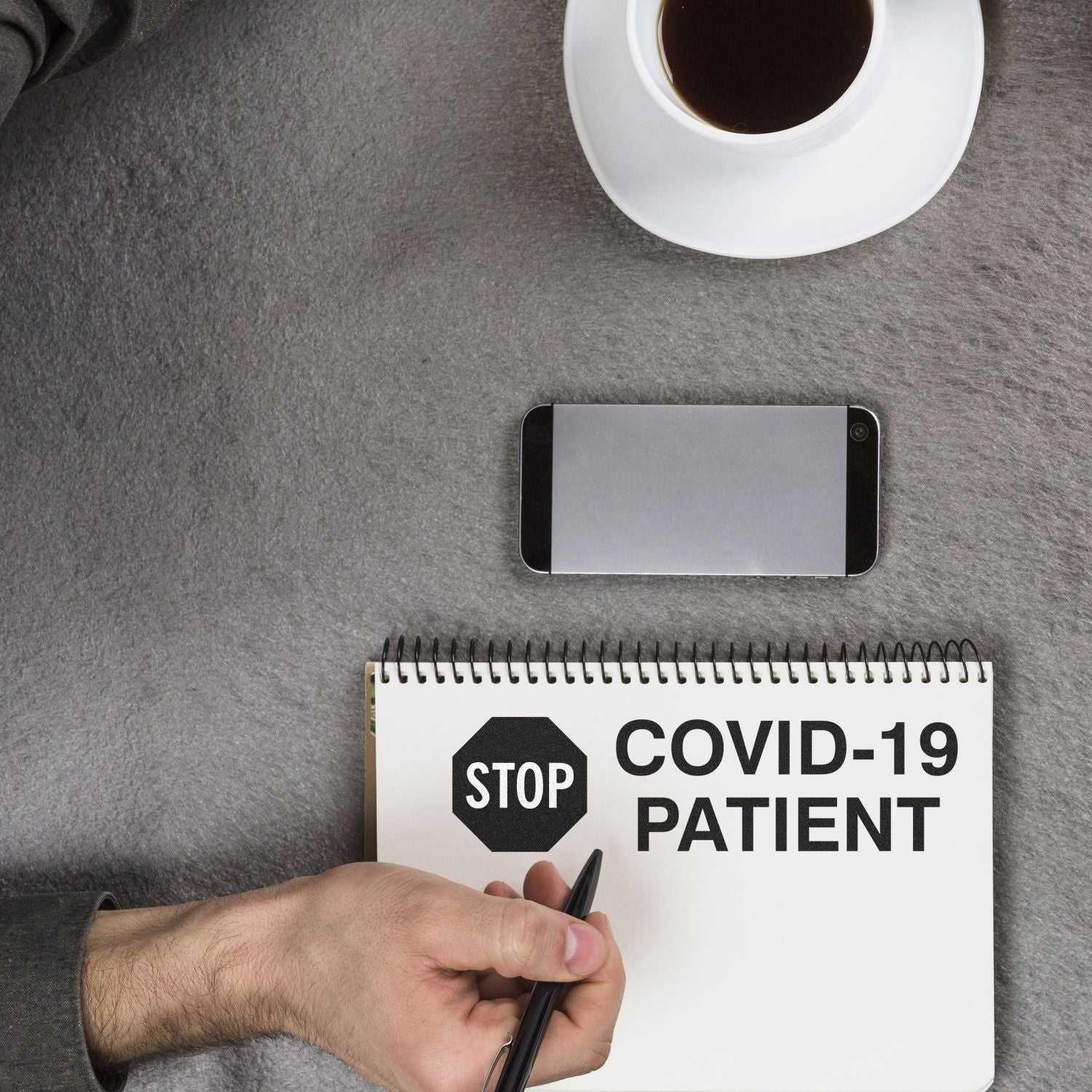 A hand holding a Large Self Inking Stop Covid Patient Stamp near a notebook with the stamp's impression, a smartphone, and a cup of coffee.