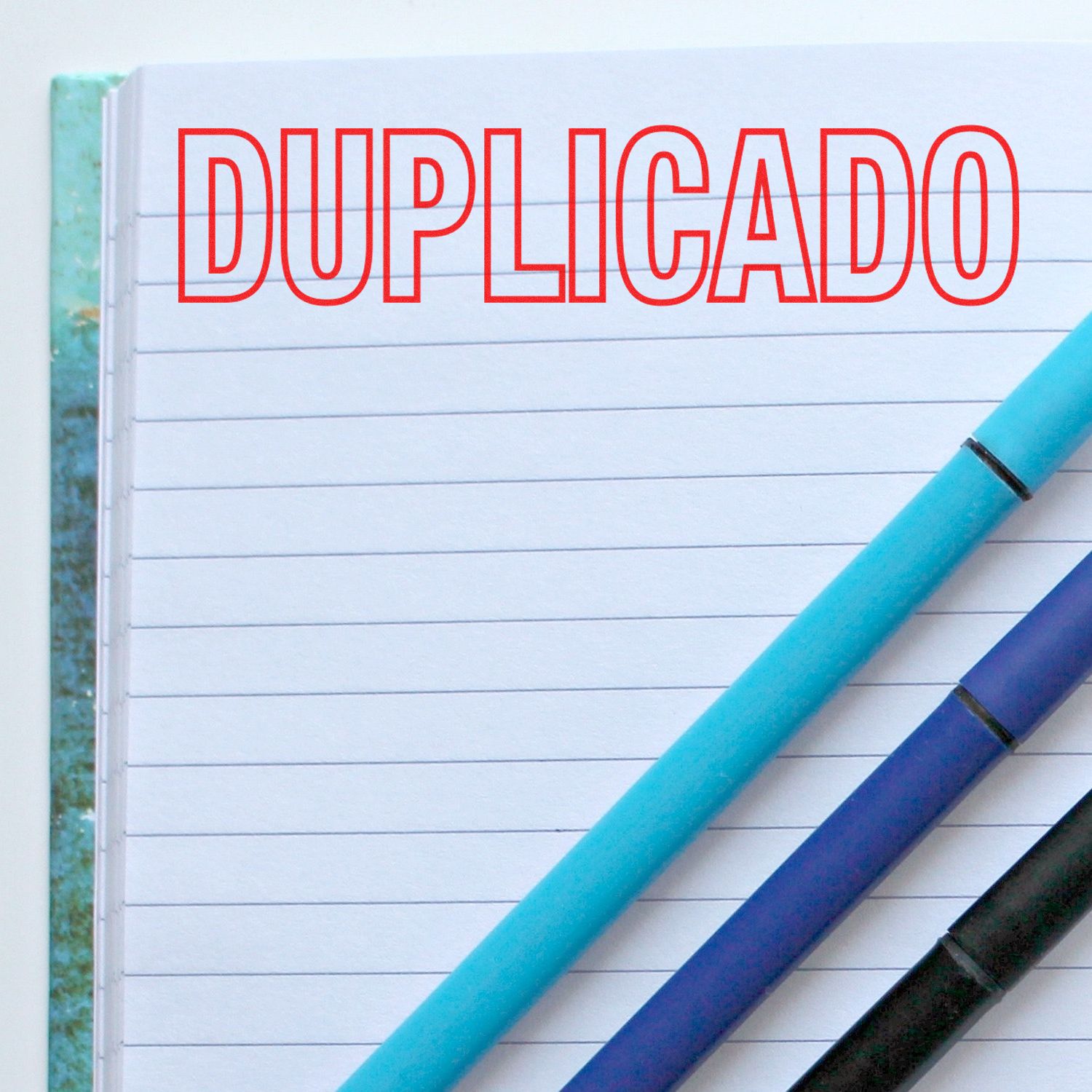 Large Self Inking Duplicado Stamp imprint on a lined notebook page with three pens placed diagonally across the page.