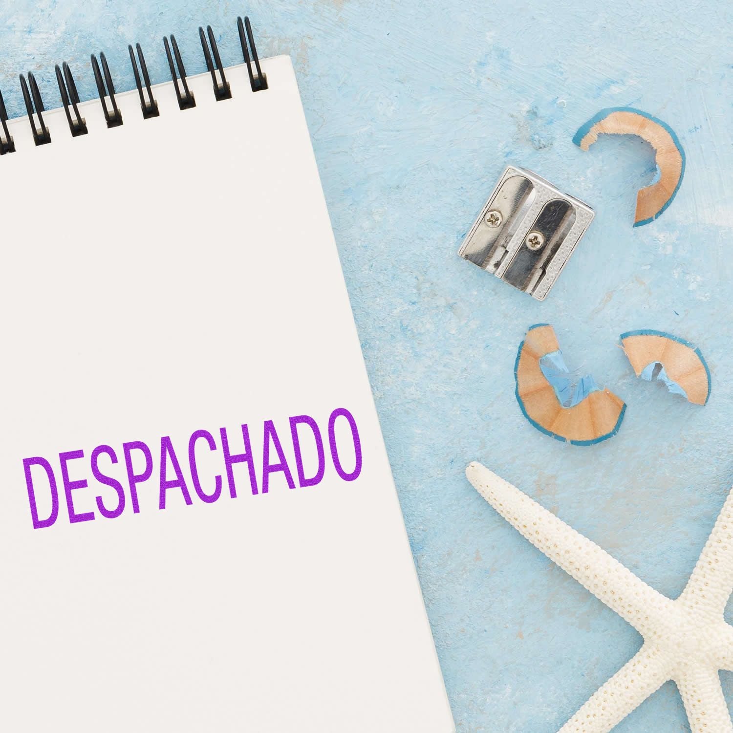 A notebook stamped with DESPACHADO using the Large Self Inking Despachado Stamp, next to a pencil sharpener, shavings, and a starfish.