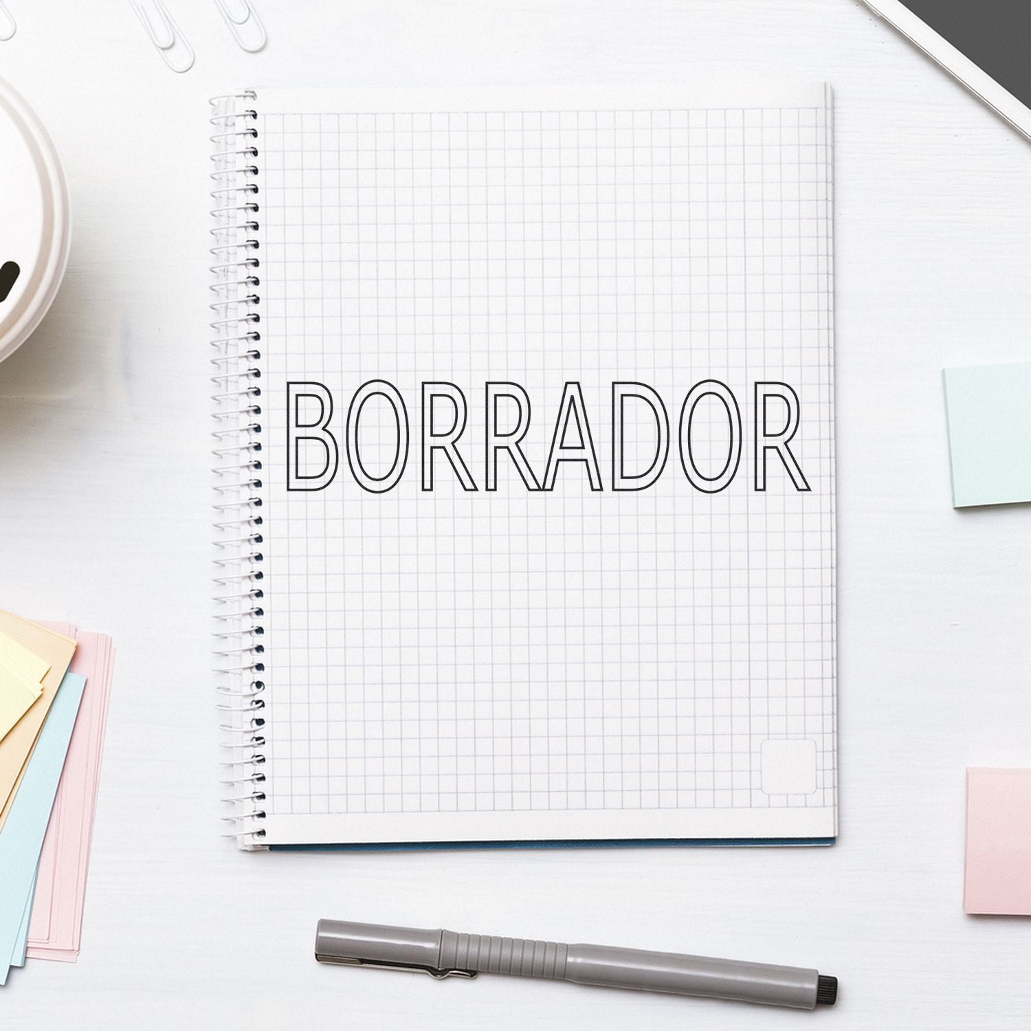 Large Self Inking Borrador Stamp on a spiral notebook with grid paper, surrounded by a pen, sticky notes, and paper clips on a white desk.