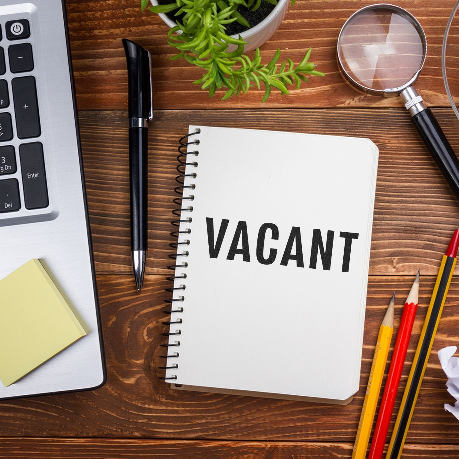 Large Pre-Inked Vacant Stamp on a spiral notebook, surrounded by office supplies including a laptop, pen, magnifying glass, and plant.