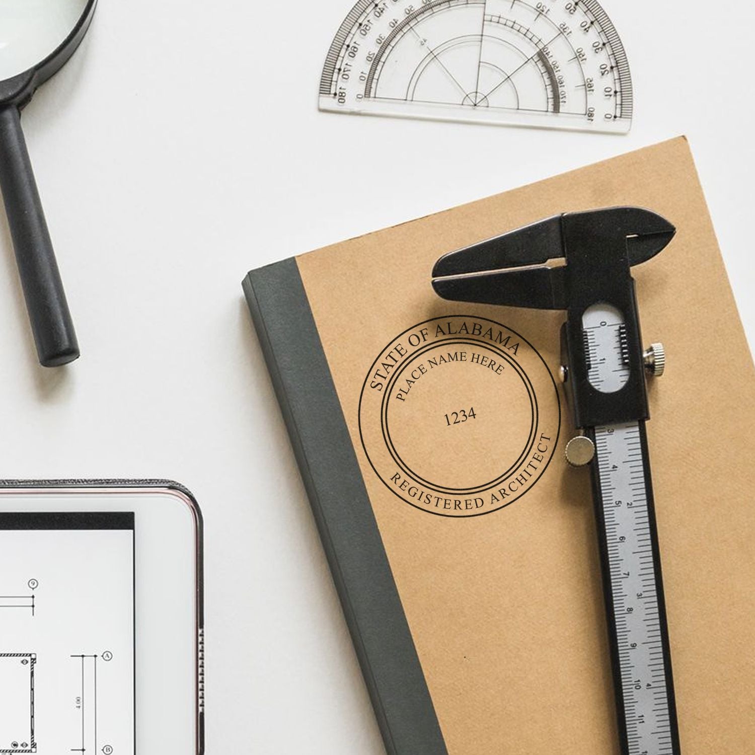 Alabama Architect Seal Stamp on a notebook with a caliper, protractor, magnifying glass, and tablet displaying architectural plans.