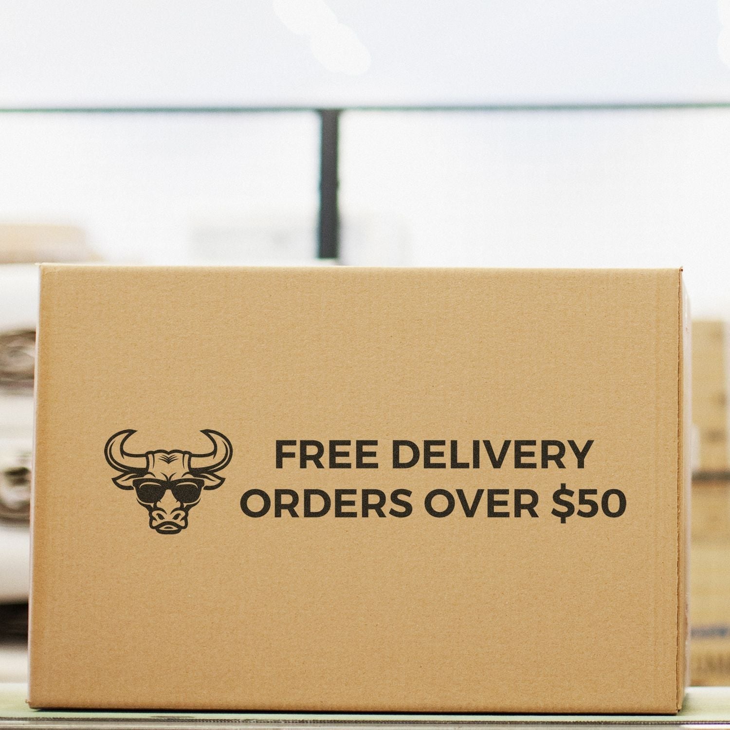 A cardboard box with FREE DELIVERY ORDERS OVER $50 stamped using a 2 Line Custom Rubber Stamp with Wood Handle, in a warehouse setting.