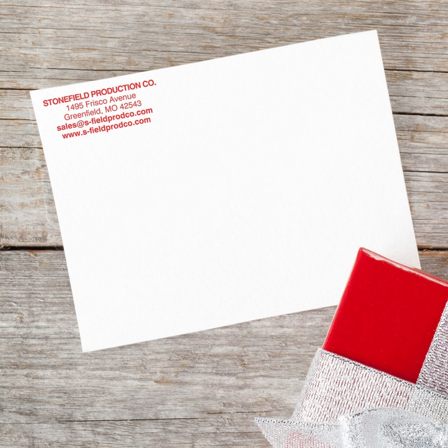 A 5 Line Custom Rubber Stamp with Wood Handle imprints red text on a white card, placed on a wooden surface next to a red gift box.