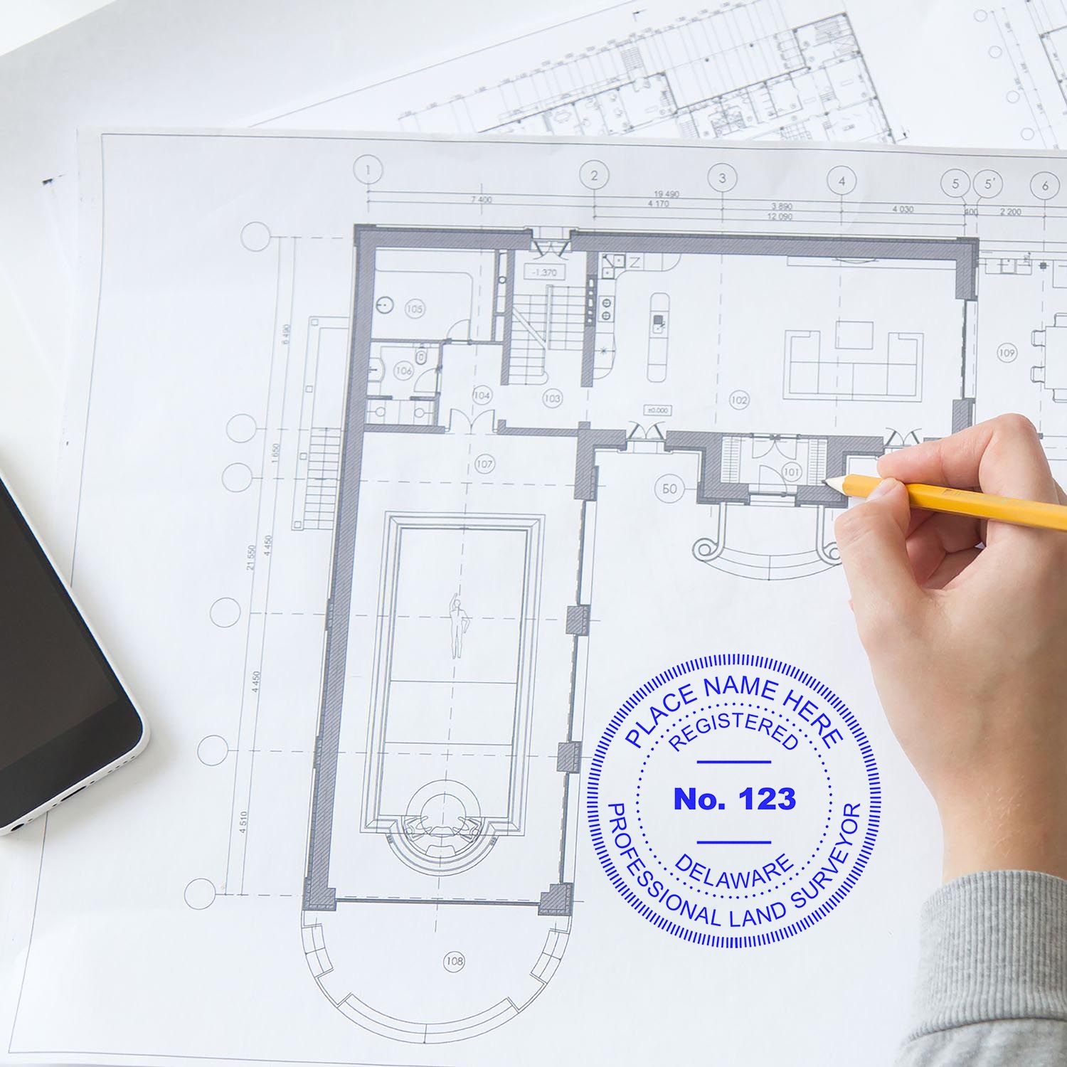 Person using the Premium MaxLight Pre-Inked Delaware Surveyors Stamp on a blueprint with a pencil and smartphone nearby.