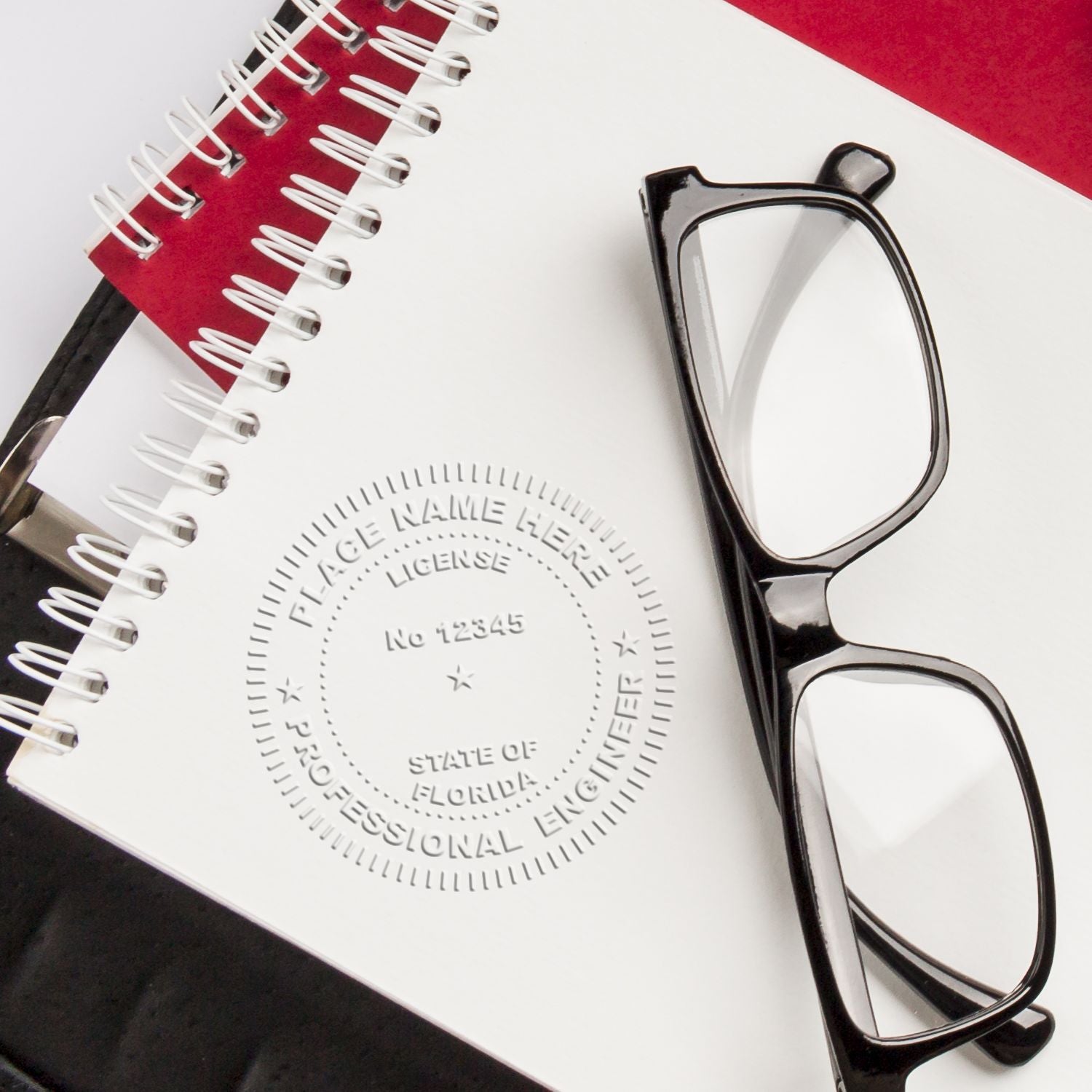 The Professional Engineer Handheld Seal Embosser is shown in use, creating a detailed embossed seal on a white notepad. The seal includes text for license number and state. Glasses and notebooks are nearby.