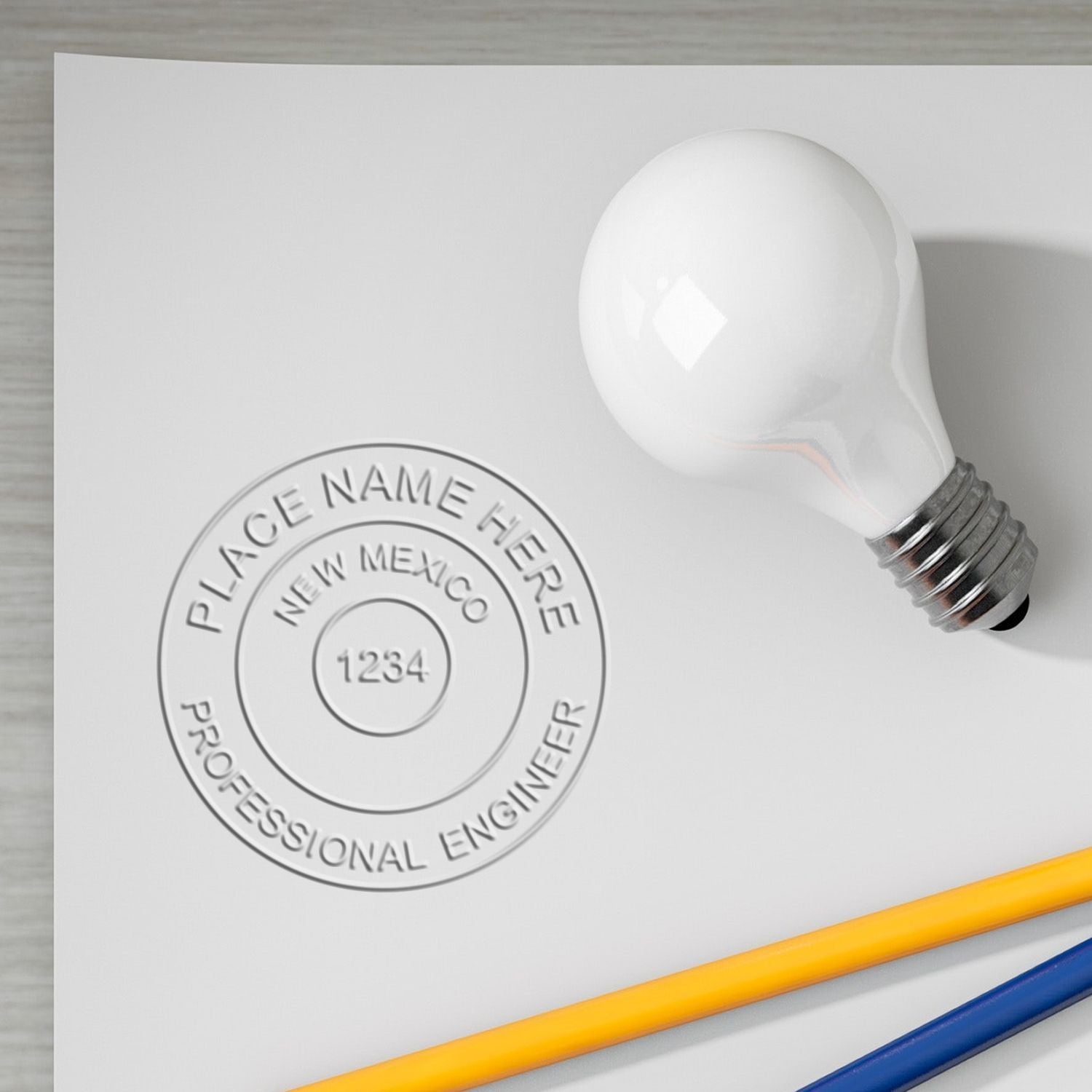 Professional Engineer Black Gift Seal Embosser imprint on white paper, showing a circular design with 'Place Name Here,' 'New Mexico,' and 'Professional Engineer.' A light bulb and pencils are nearby.