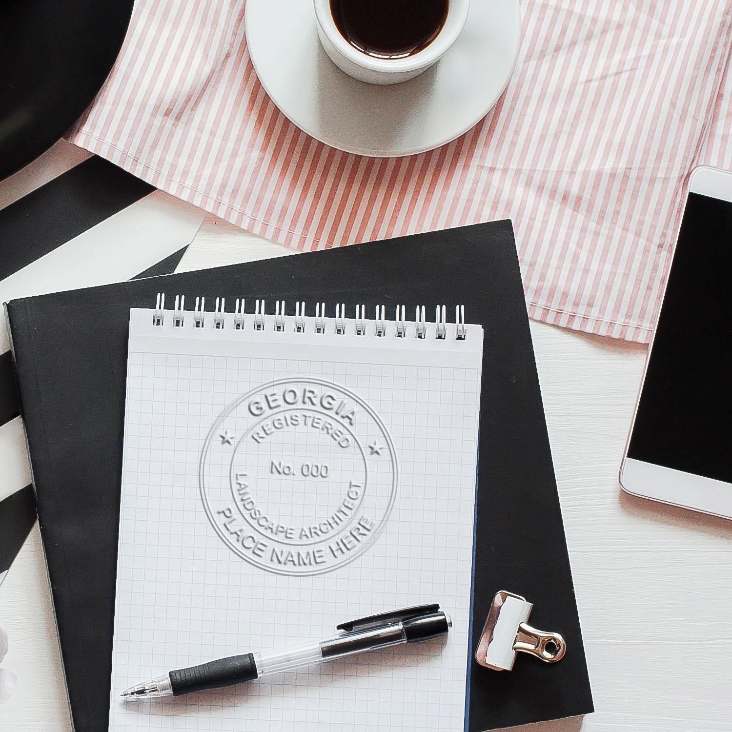 Landscape Architect Black Gift Seal Embosser in use on a notepad, with a pen, clipboard, coffee cup, and phone on a white desk.