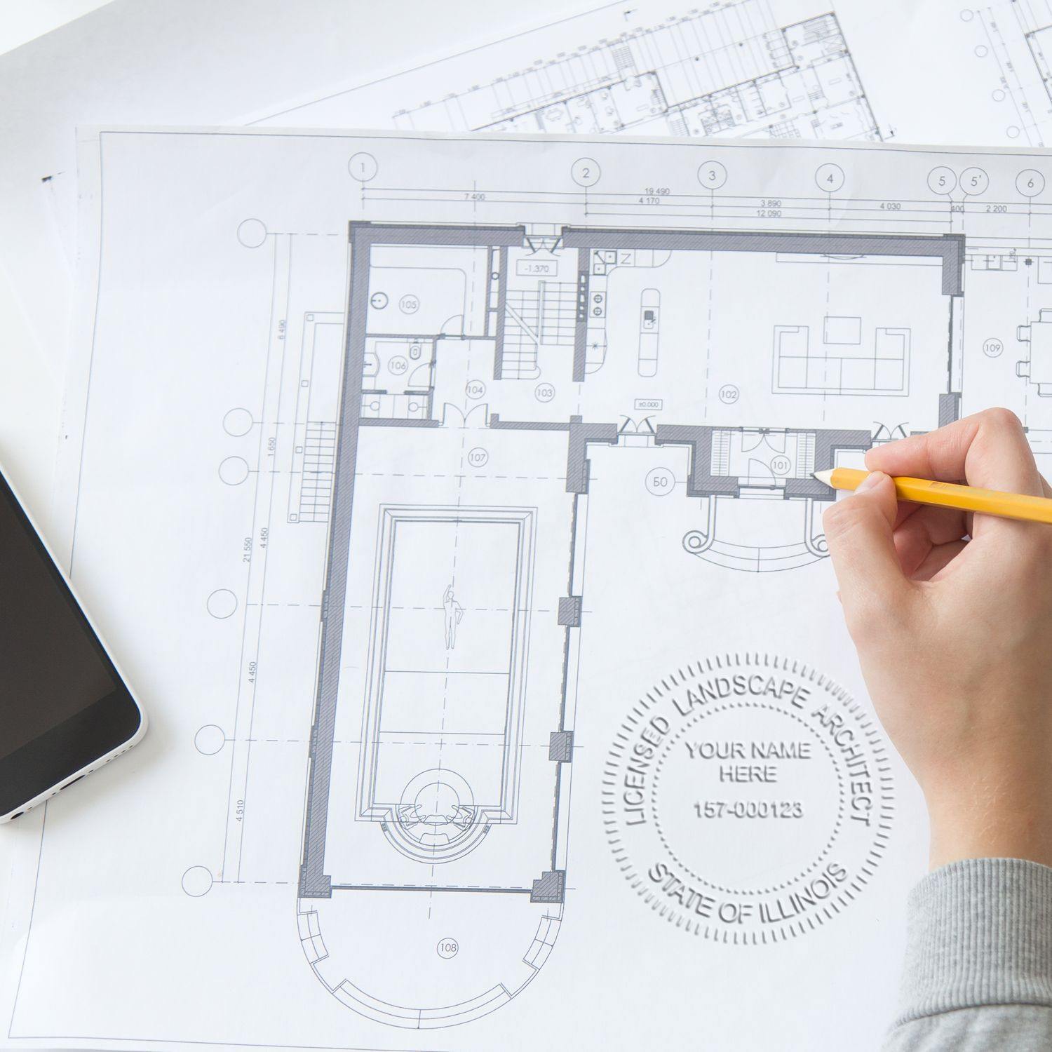 Hand using the Landscape Architect Long Reach Desk Seal Embosser on architectural blueprints, with a smartphone nearby.