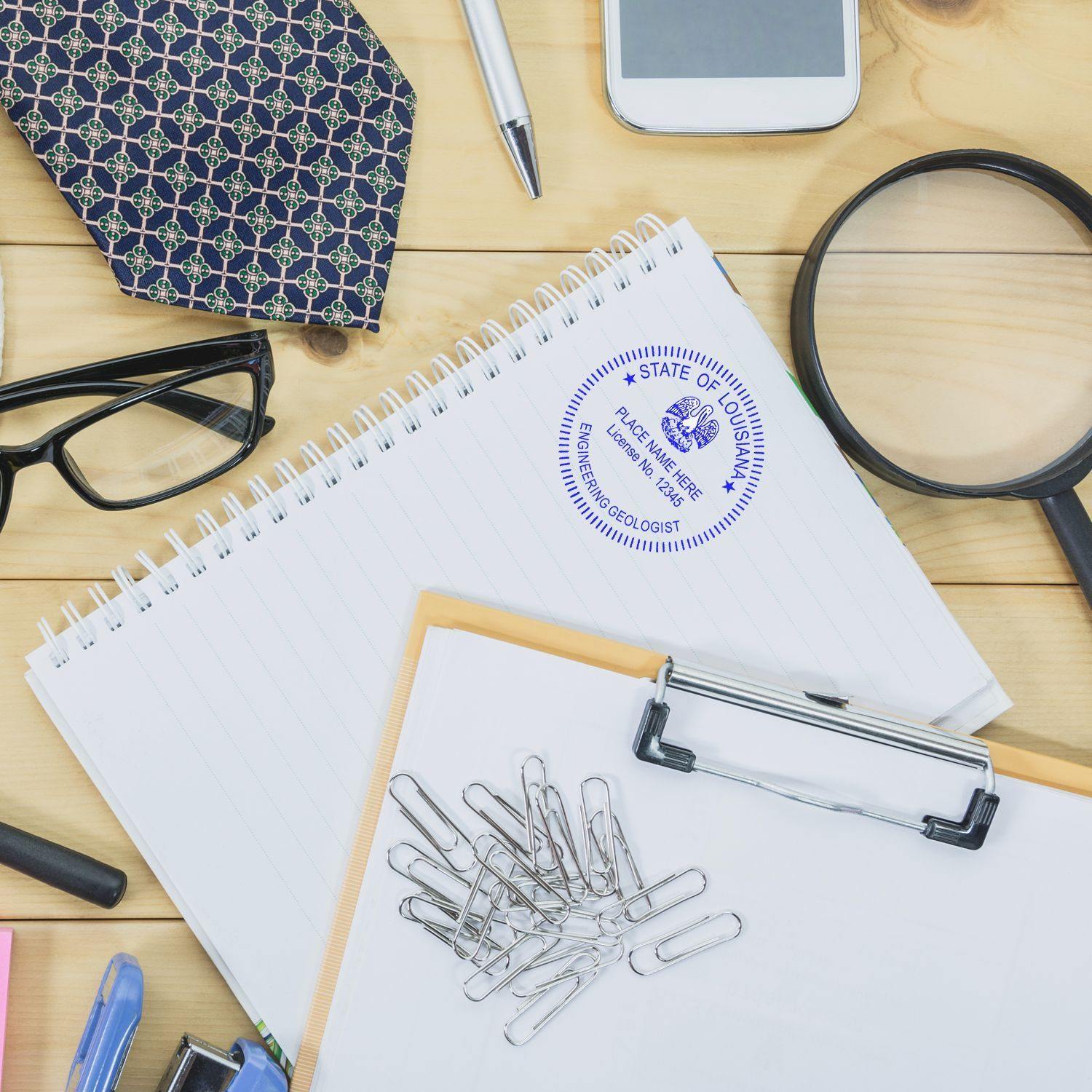 A desk with office supplies, including a notepad stamped with the Engineering Geologist Regular Rubber Stamp of Seal, a magnifying glass, paper clips, glasses, a tie, and a smartphone.