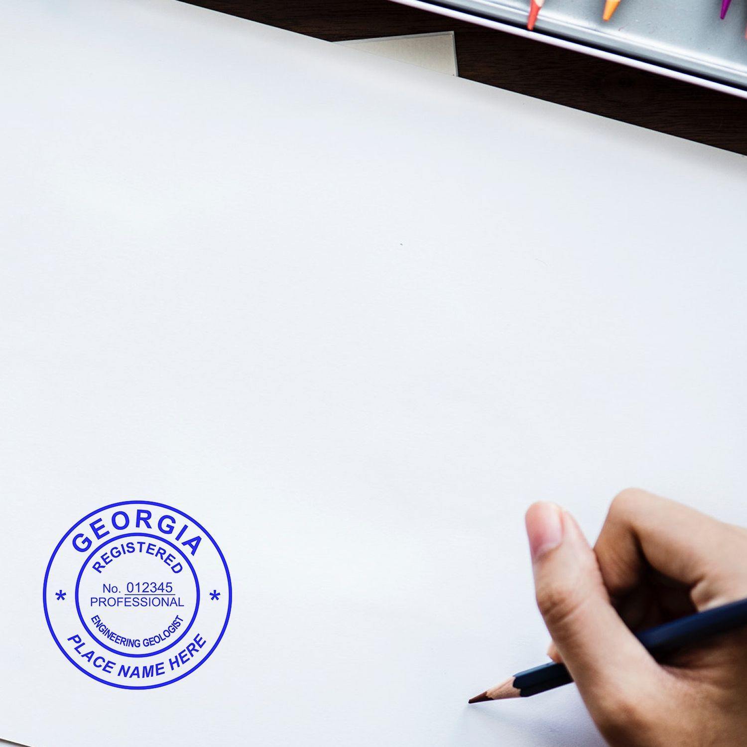 A hand holding a pencil near a stamped paper with the Engineering Geologist Regular Rubber Stamp of Seal, featuring a blue circular design with Georgia Registered Professional Engineering Geologist text.