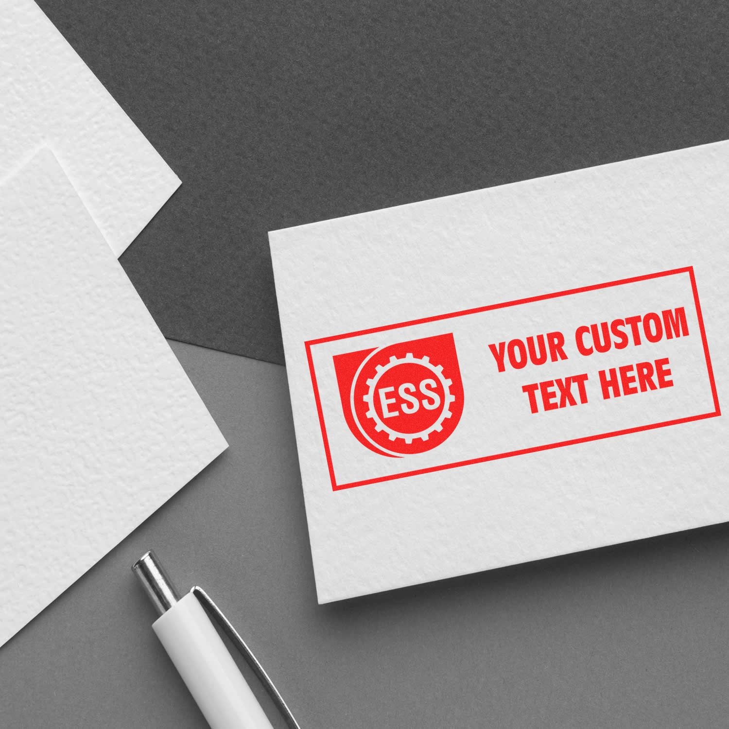 Custom Rubber Stamp Size 1 x 2 in red ink on a white card with ESS logo and Your Custom Text Here beside a white pen on a gray surface.