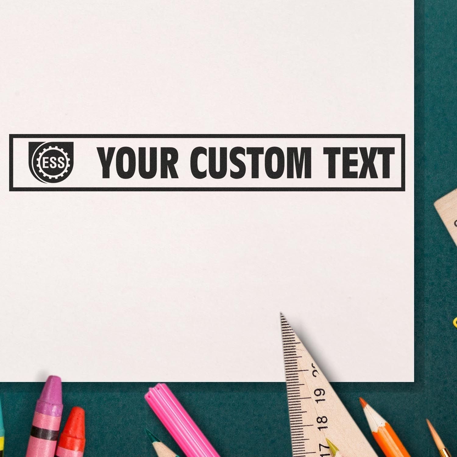 Custom Rubber Stamp Size 1 x 10 imprinting YOUR CUSTOM TEXT on paper, surrounded by colorful pencils and a ruler on a green surface.