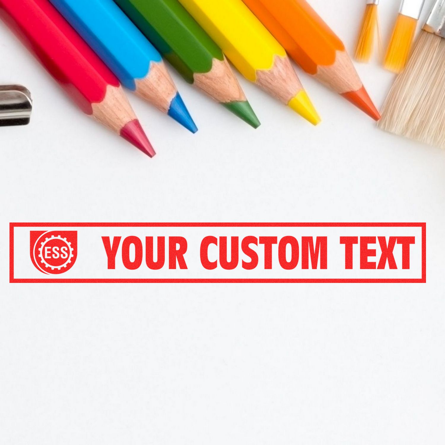 Custom Rubber Stamp Size 2 x 7 in use, displaying YOUR CUSTOM TEXT in red ink, surrounded by colorful pencils and paintbrushes.