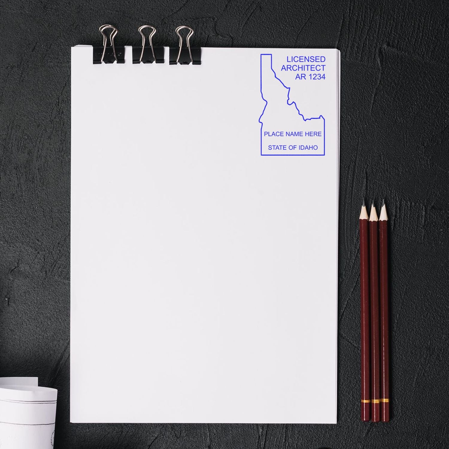 Self Inking Idaho Architect Stamp imprint on a white paper with three pencils and binder clips on a dark surface.