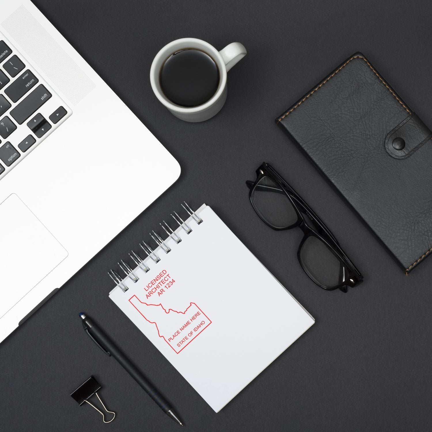 A Premium MaxLight Pre-Inked Idaho Architectural Stamp on a notepad, surrounded by a laptop, coffee cup, glasses, wallet, pen, and binder clip.