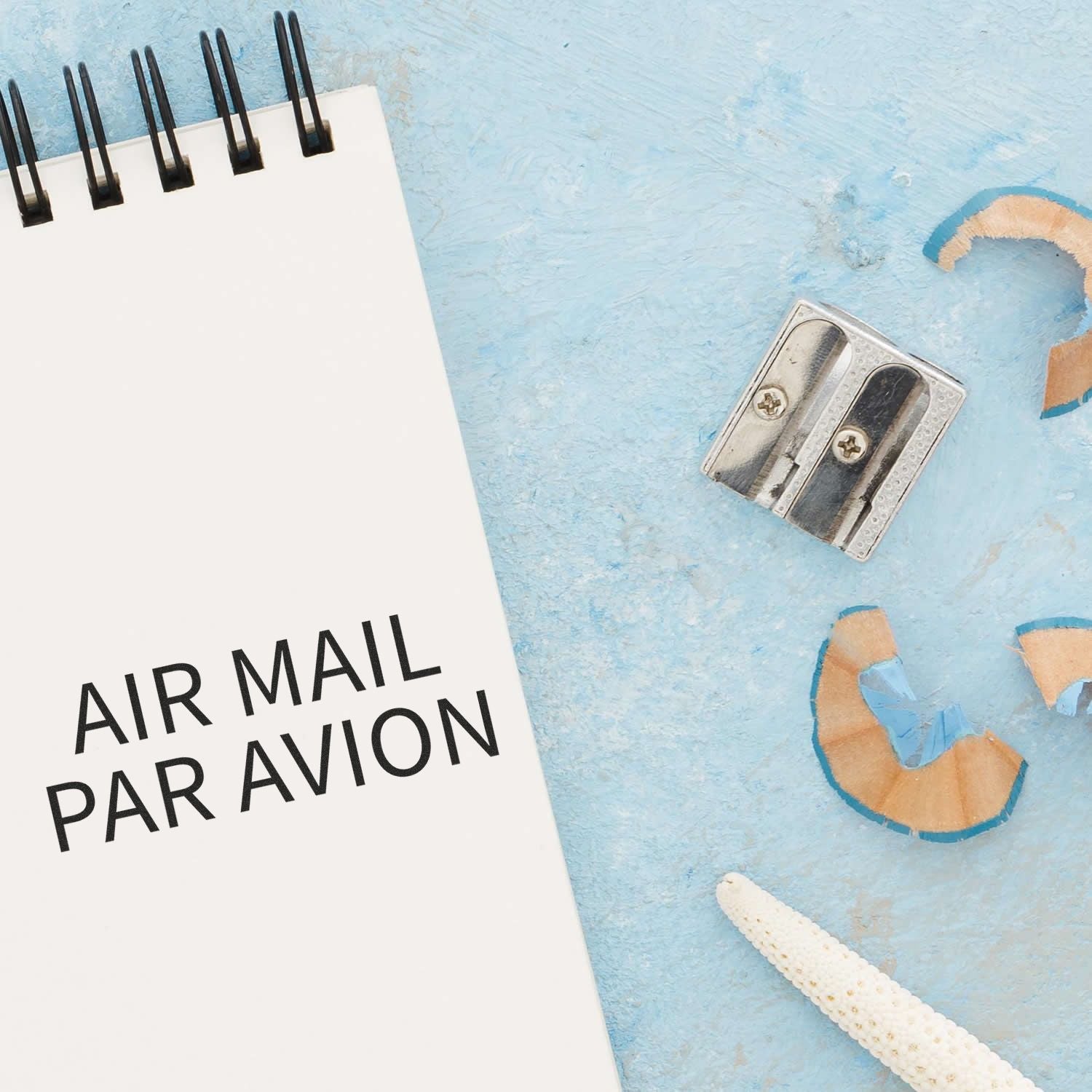 Self Inking Air Mail Par Avion Stamp used on a notebook, next to a pencil sharpener and shavings on a light blue surface.