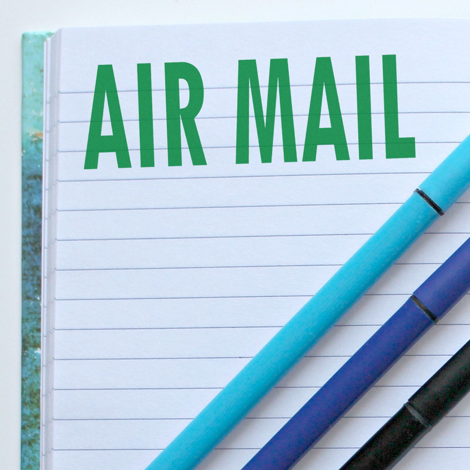 Self Inking Air Mail Stamp used on a lined notebook page with AIR MAIL in green letters, next to two blue pens.