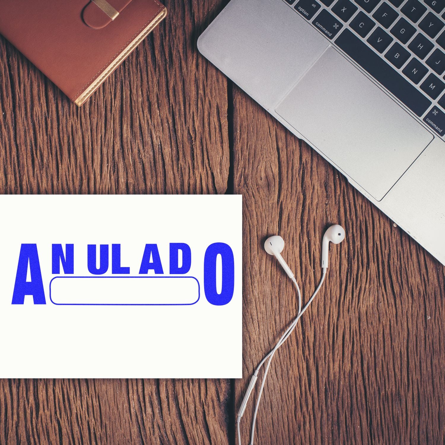 Large Anulado Rubber Stamp in blue ink on paper, placed on a wooden desk next to a laptop, earphones, and a brown notebook.