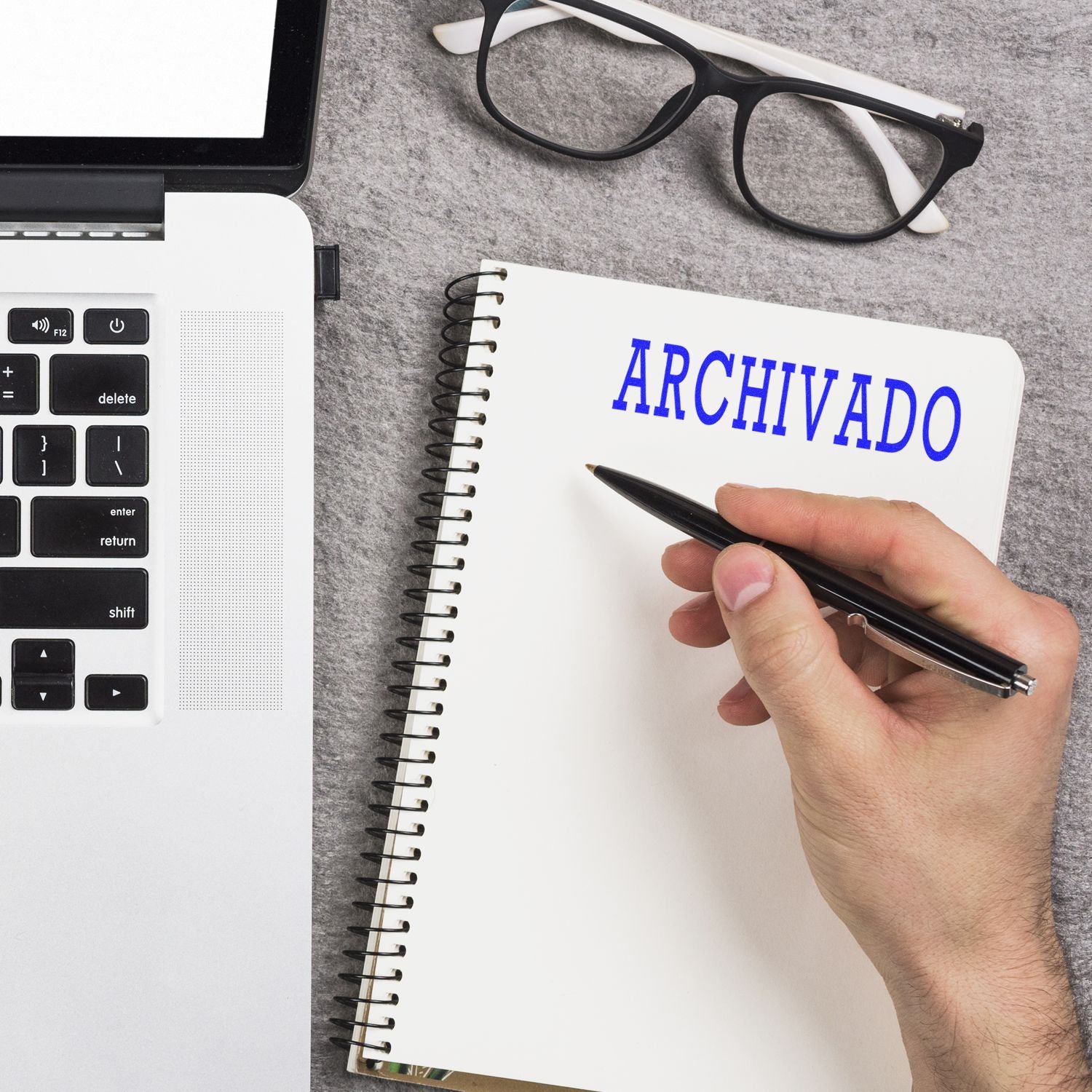 Hand holding pen near notebook with Archivado rubber stamp imprint, next to a laptop and glasses on a gray surface.