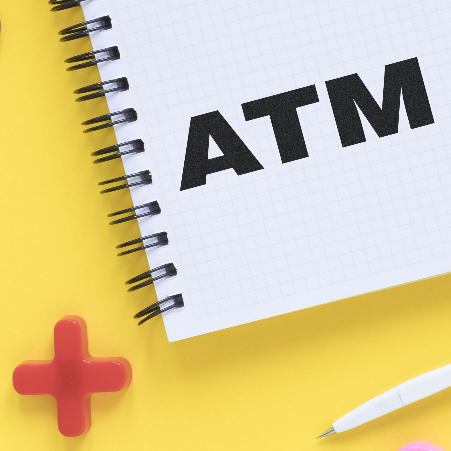 Notebook with ATM text on a yellow background, next to a red cross symbol and a white pen. Focus: Bank Stamp ATM Rubber Stamp.