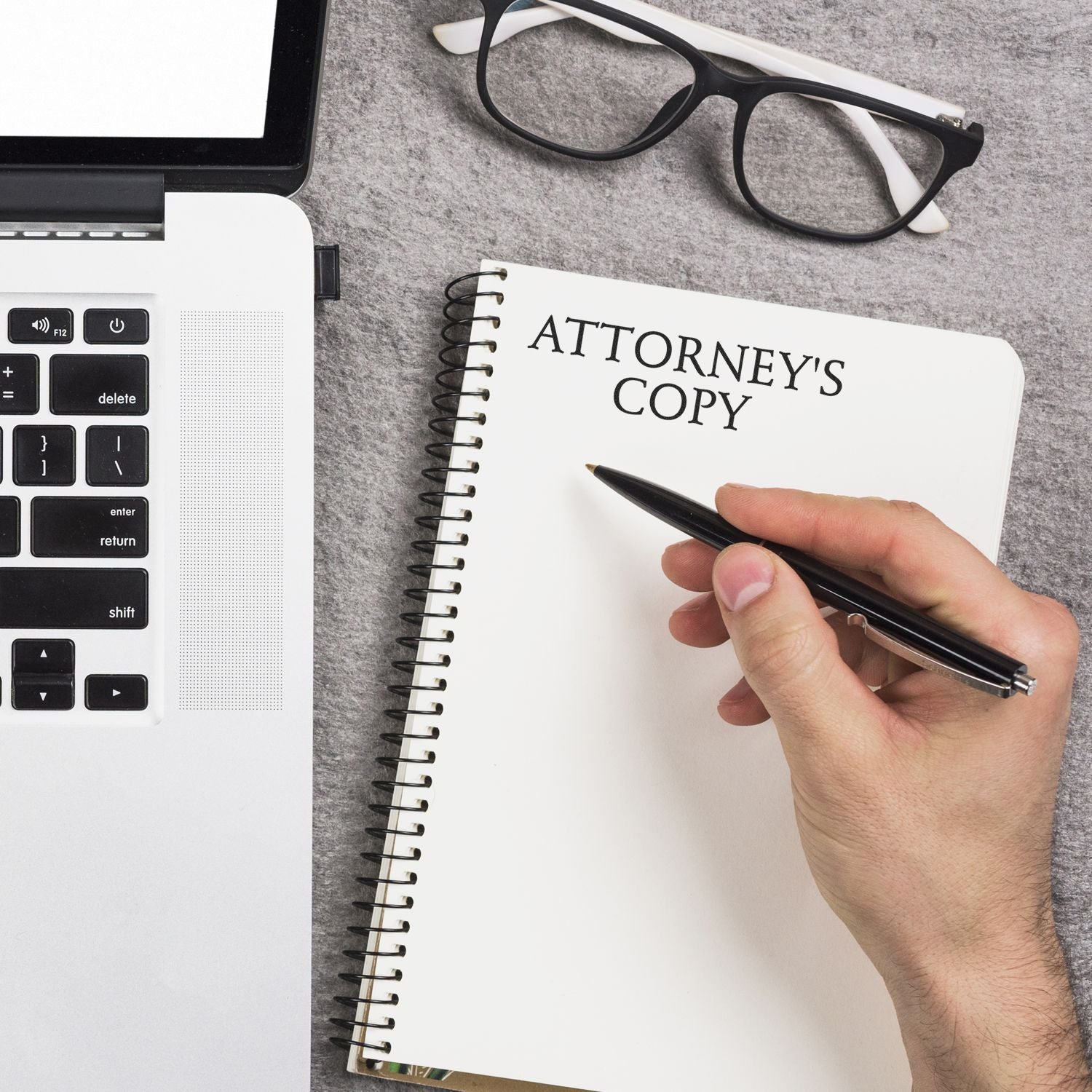 Hand writing in a notebook labeled ATTORNEY'S COPY next to a laptop and glasses, with an Attorneys Copy Rubber Stamp in use.