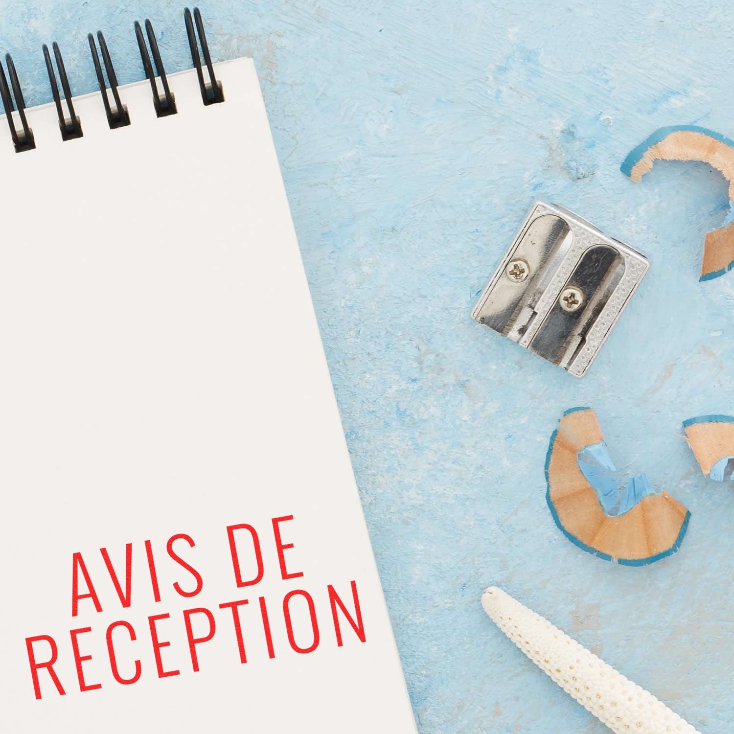 Large Pre-Inked Avis De Reception Stamp used on a notepad, with a pencil sharpener and shavings nearby on a light blue surface.