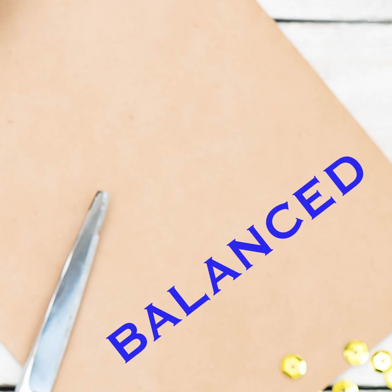 Large Balanced Rubber Stamp imprinting the word BALANCED in blue ink on a beige surface, with scissors and sequins nearby.