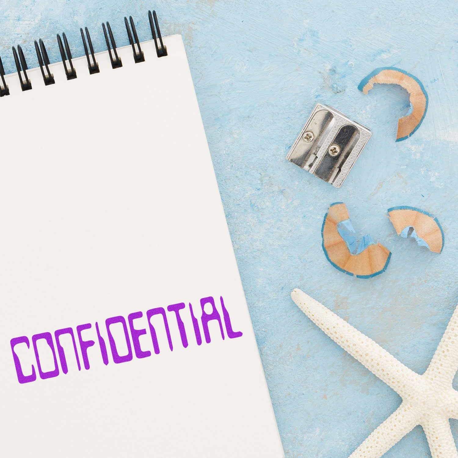 A notebook stamped with CONFIDENTIAL using the Large Barcode Confidential Rubber Stamp, next to a pencil sharpener and starfish on a blue surface.