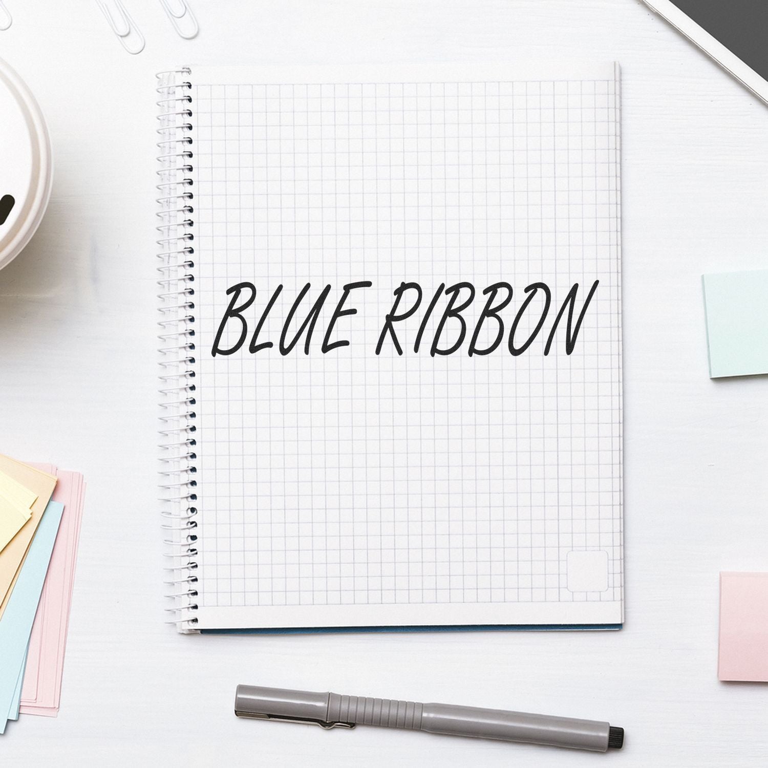 Notebook with 'Blue Ribbon' stamped on grid paper, surrounded by a pen, sticky notes, and paper clips.