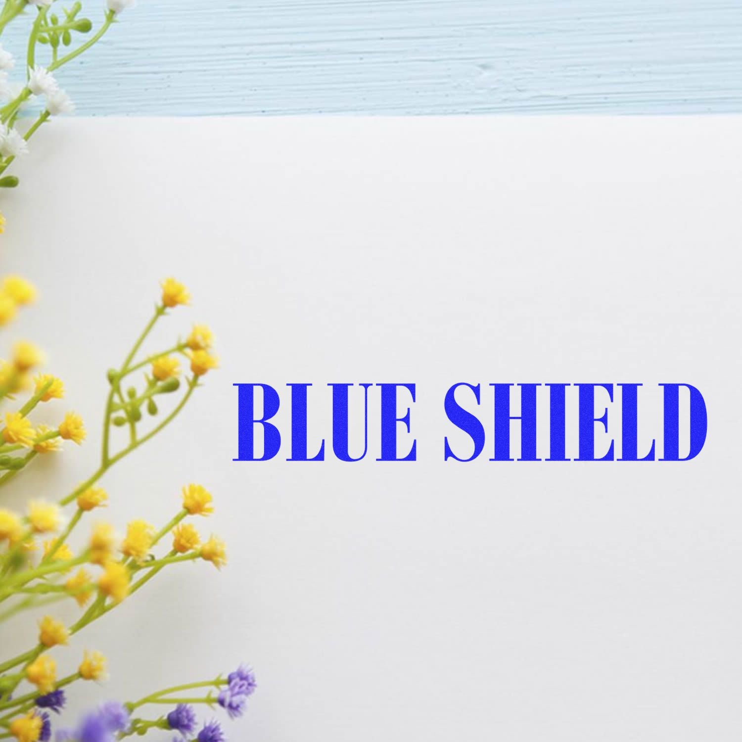 Blue Shield rubber stamp impression on white paper with yellow and purple flowers on the side.